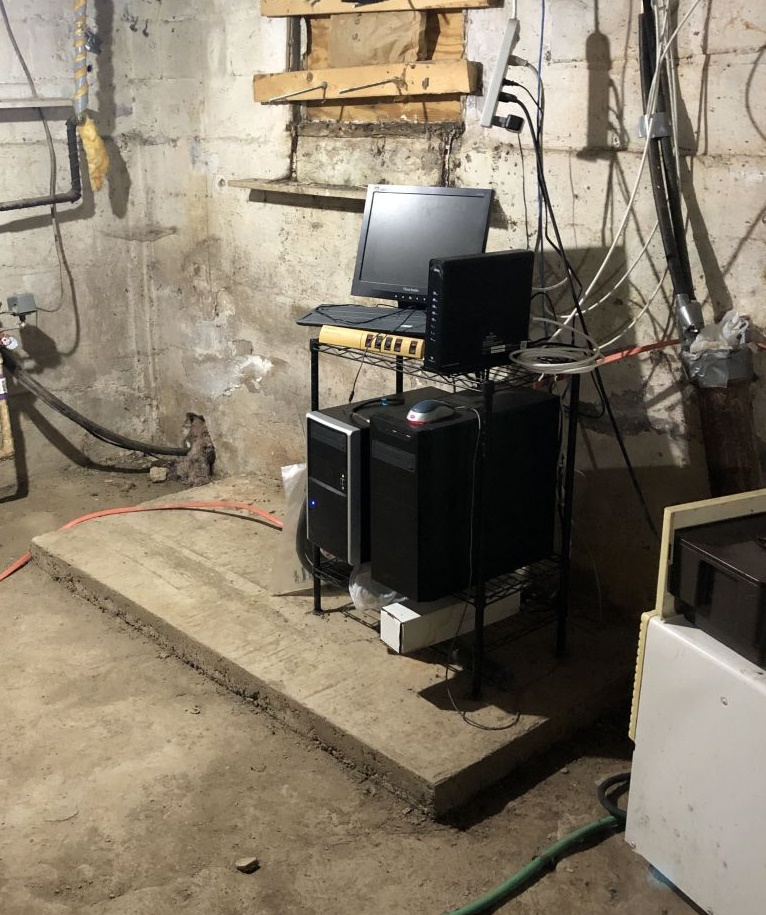 Monitor, servers, and a modem are resting on a short metal shelf in a cobweb filled, clearly damp, grungy basement.
